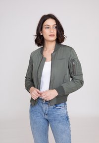 Olivengrøn bomberjakke med ribbede manchetter og kant, lynlåslomme på ærmet og glat tekstur. Kombineret med højtaljede lyseblå jeans.