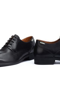 Paar schwarze Leder-Herrenschuhe mit Schnürsenkeln, niedrigen Absätzen und „Pikolinos“-Markenlabels an den Seiten, vor weißem Hintergrund gezeigt.