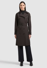 Sieviete, kura valkā tumši brūnu dubultrindu trenci ar jostu, ceļgala garuma, melnas platajām biksēm un melnus zābakus, stāv taisni.