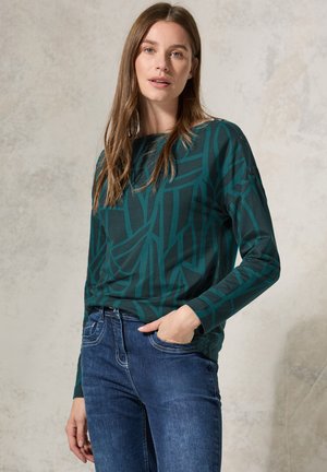 Chemise verte à manches longues avec un imprimé géométrique, fabriquée en matériau doux. Associée à un jean bleu. Présente une coupe décontractée et un col rond.