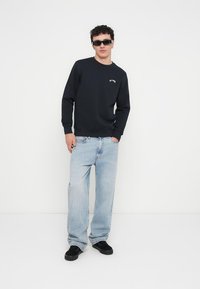 Fekete pulóver kerek nyakkal és kis emblémával, világoskék bő nadrággal és fekete tornacipővel párosítva. A modell napszemüveget visel.