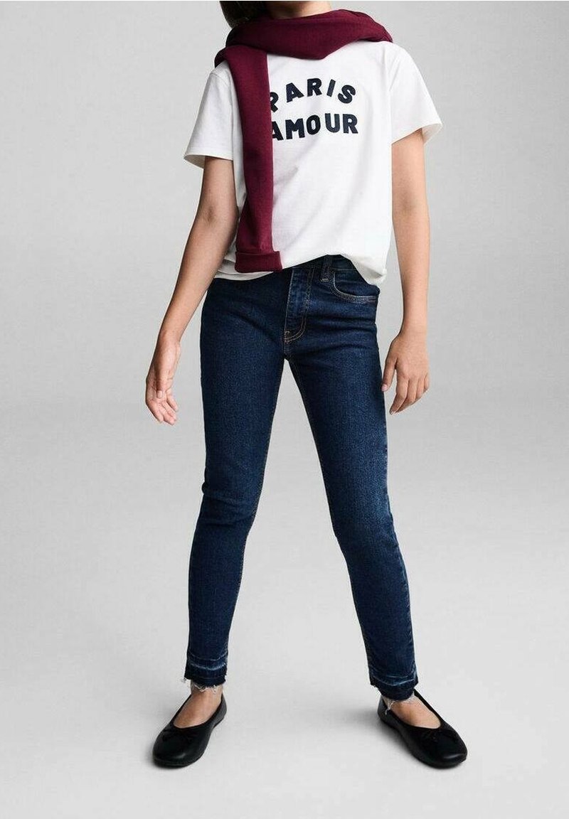 Camisa blanca con texto negro "PARIS AMOUR", jeans oscuros y zapatos planos negros. Un suéter burdeos está drapeado sobre los hombros.