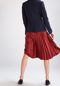 Marineblazer kombiniert mit einem plissierten, knielangen, burgunderfarbenen Rock. Der Rock hat eine glatte, glänzende Textur. Schwarze Schuhe runden das Outfit ab.