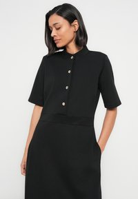 Μαύρο φόρεμα με κοντά μανίκια και όρθιο γιακά, διακοσμημένο με μια σειρά ασημένιων κουμπιών κατά μήκος του μπροστινού μέρους και πλαϊνές τσέπες. Λείο υφασμάτινο υλικό.