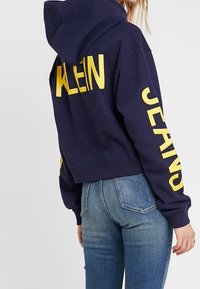 Person som bär en marinblå hoodie med gul "KLEIN" på ryggen och "JEANS" på ärmen, ihopparat med blå denimjeans, visad bakifrån.
