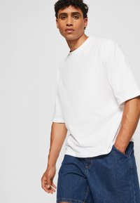 Tricou alb din bumbac, cu guler rotund și mâneci scurte, asortat cu pantaloni scurți din denim albastru, care au un buzunar și detalii cusute vizibile.