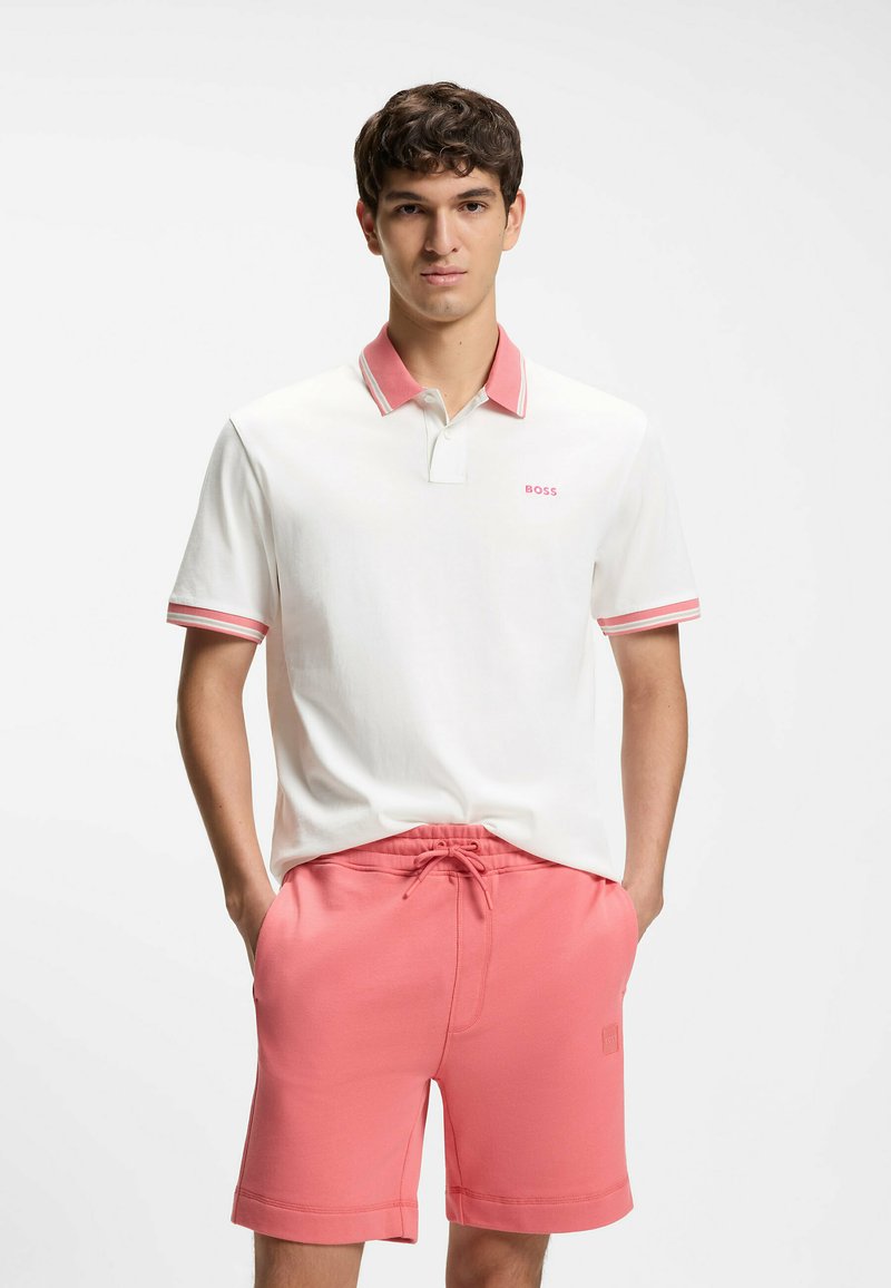 Jeune homme debout les mains dans les poches, portant un polo blanc avec un col rose et un short rose assorti sur fond blanc.