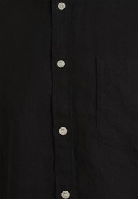 Devant de chemise en tissu noir avec boutons blancs et poche poitrine, montrant la patte de boutonnage et le matériau texturé.