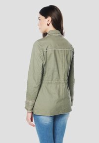 Olivo zelena jakna z vezenimi poudarki na rokavih, detajlom z zbiranjem v pasu in klasičnim ovratnikom. Oblečena z modrimi kavbojkami.