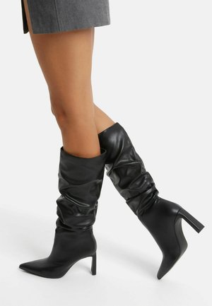 Ben iført sorte læderstøvler med hæl og spidse tæer, der går til midt på læggen, kombineret med en kort grå nederdel.