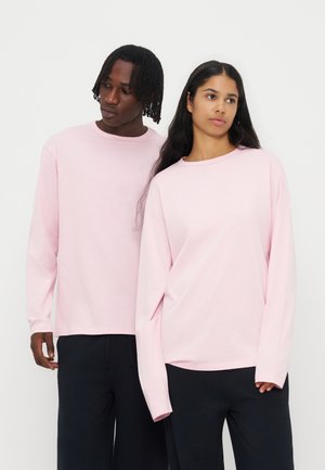 Deux adultes portant des chemises à manches longues rose clair assorties et des pantalons noirs se tenant devant un fond blanc.