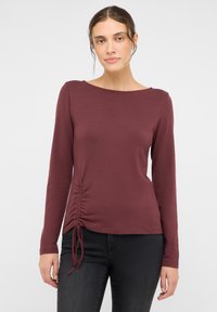 Bordeauxrotes Langarmshirt mit einem Bootsausschnitt und seitlicher Raffung. Glatter Stoff, lässige Passform, kombiniert mit dunklen Jeans.