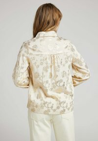 Blouse en satin doré avec un motif floral, dotée d'un col, d'un dos froncé et de manches longues, assortie à un pantalon blanc. Texture lisse, léger éclat.