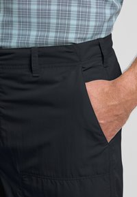 Pantaloni neri leggeri realizzati in tessuto liscio. Presentano una tasca laterale e dettagli di cucitura discreti. Abbinati a una camicia a quadri blu e grigia.