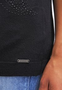 Maglione nero a maglia con un motivo testurizzato di perline nere e un'etichetta argentata con il marchio lungo l'orlo inferiore. Sfondo in denim chiaro.