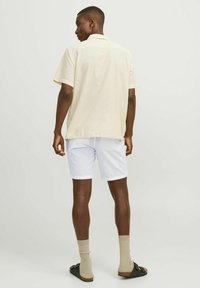 Camisa corta de manga corta en beige claro con un diseño texturizado, combinada con pantalones cortos blancos. El atuendo se complementa con calcetines beige y sandalias negras.