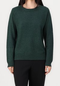Femme portant un pull à manches longues vert foncé à col rond et un pantalon noir, debout devant un fond blanc.
