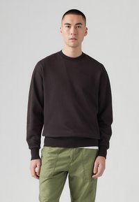 Sudadera marrón con cuello redondo y puños de canalé, combinada con pantalones verdes oliva, presenta detalles sutiles del logo. Textura suave.