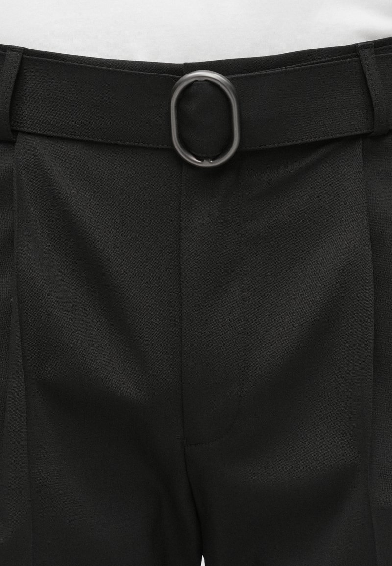 Pantaloni neri a vita piatta, con passanti larghi e fibbia metallica rettangolare. Il tessuto sembra liscio e strutturato.