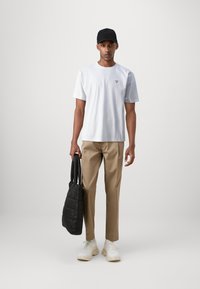 Hellblaue Baumwoll-T-Shirt mit kleinem roten dreieckigen Logo, beigen Hosen, schwarzer Kappe, hält eine gesteppten schwarzen Tote-Bag und weiße Sport Schuhe.