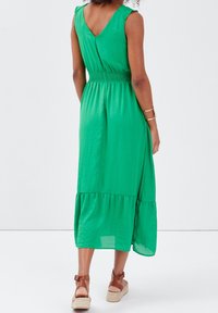 Robe verte sans manches avec une taille smockée, un ourlet à volants et un dos en V. Portée avec des sandales espadrilles couleur camel, le tissu semble léger.