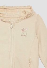Giacca con cappuccio beige chiaro con zip. Materiale morbido con ricami floreali e orlo arricciato lungo il cappuccio. La zip presenta un design a forma di fiore.