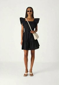 Vestido preto com mangas em babados, corpete ajustado e saia em camadas. Combinado com uma bolsa creme e sandálias brancas. Tecido suave, design casual.