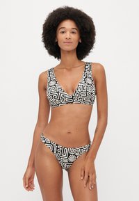 Čierny a biely bikiny set so geometrickým vzorom. Vrchná časť má široké ramienka a hlboký výstrih; spodná časť sú nízko vyrezané s rovnakým dizajnom.