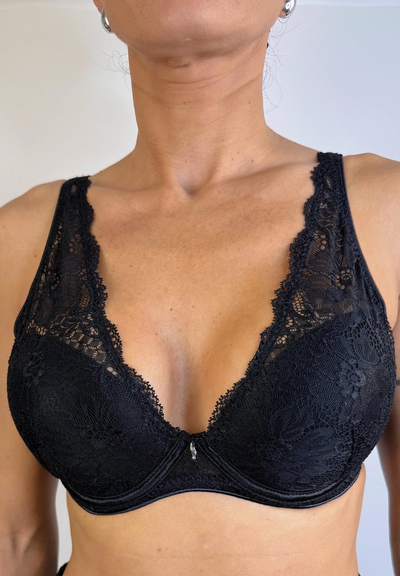 Sujetador de encaje negro con un profundo escote en V, bordes festoneados y un pequeño adorno decorativo en el centro. Tela suave debajo del encaje.