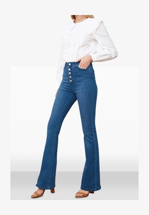 Jean évasé en denim bleu taille haute avec fermeture à cinq boutons et deux poches avant, porté avec un chemisier blanc à manches longues.