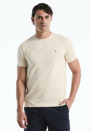 Mann mit dunklen lockigen Haaren, der ein beigefarbenes kurzärmeliges T-Shirt und dunkle Hosen trägt, vor einem schlichten weißen Hintergrund steht.