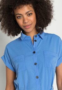 Chemise bleue à manches courtes retroussées, avec deux poches poitrine et des boutons noirs, fabriquée dans un tissu lisse avec un léger éclat.