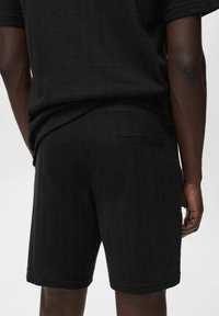 Vue arrière d'une personne portant un short noir texturé et un haut noir à manches courtes assorti, devant un fond uni.
