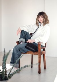 Černá krosová taška z hladké kůže, nošená s uvolněnou bílou blůzou a vybledlými džíny. V pozadí dřevěná židle a zelená rostlina.