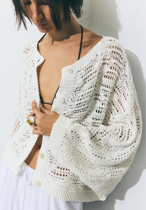 Kvinde iført en hvid åbenstrikket cardigan over en sort bikini top, der holder cardigans lukket, med en stor ravring på fingeren.