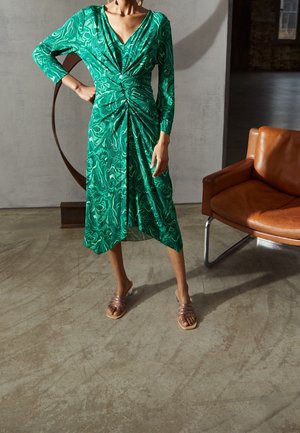 Kobieta ubrana w zieloną, wzorzystą sukienkę midi z długim rękawem, zawiązaną węzłem w talii, stojąca obok brązowego skórzanego fotela na betonowej podłodze.