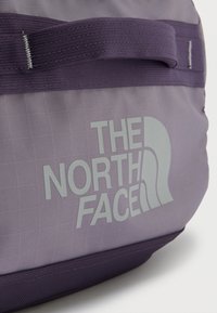 Κοντινό πλάνο μοβ τσάντας The North Face που δείχνει το λογότυπο και τη ραμμένη λαβή πάνω σε ύφασμα με διακριτικό σχέδιο σε πλέγμα.