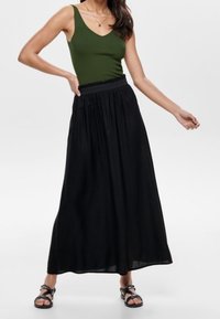 Sort maxinederdel med en rynket talje, kombineret med en tætsiddende grøn tanktop. Modellen har flade sorte sandaler på og udstråler en afslappet stil.