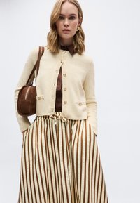 Γυναίκα που φοράει κρεμ πλεκτή ζακέτα πάνω από καφέ πουκάμισο, καφέ και κρεμ παντελόνι με κάθετες ρίγες και φαρδιά πόδια, κρατώντας καφέ σουέντ τσάντα ώμου.