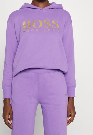 Osoba ubrana w fioletową bluzę z kapturem i dopasowane spodnie dresowe z złotym logo "BOSS HUGO BOSS" na piersi i spodniach.