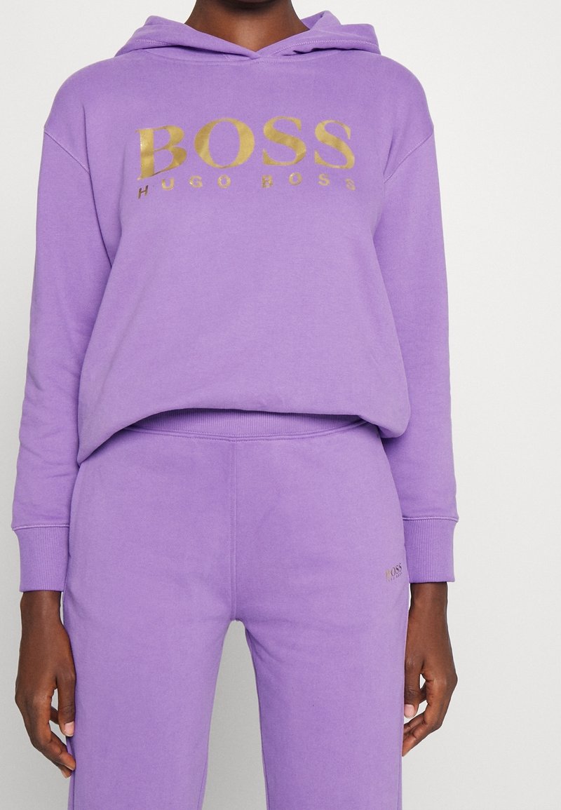 Personne portant un sweat à capuche violet et un pantalon de survêtement assorti avec un logo doré "BOSS HUGO BOSS" sur la poitrine et le pantalon.