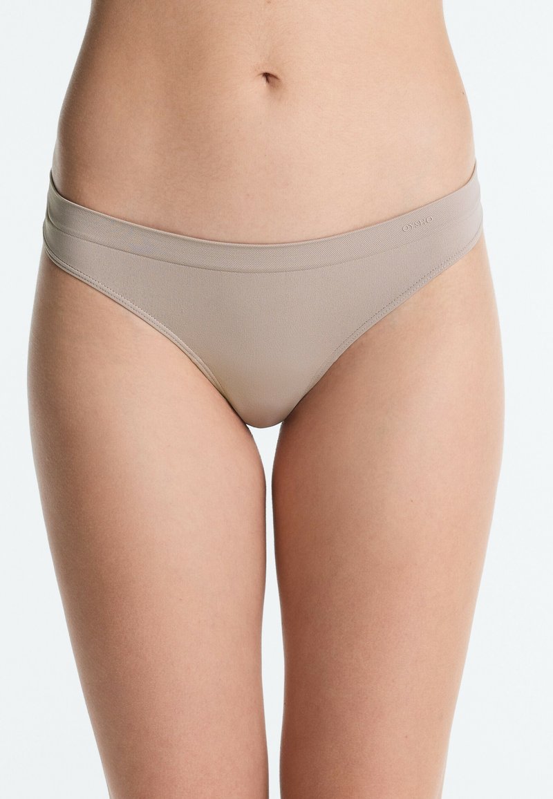 Partie inférieure du torse d'une femme portant une culotte bikini beige sans coutures sur fond blanc.