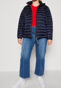 Marineblaue Steppjacke mit horizontalen Steppnähten, gefütterter Kapuze, Frontreißverschluss und Logodetail. Wird über einem roten Hemd getragen und mit hellblauen Jeans kombiniert.