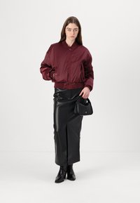 Giubbotto bomber bordeaux con polsini a coste, abbinato a una gonna di pelle nera con una fessura frontale. La modella tiene una piccola borsa nera.