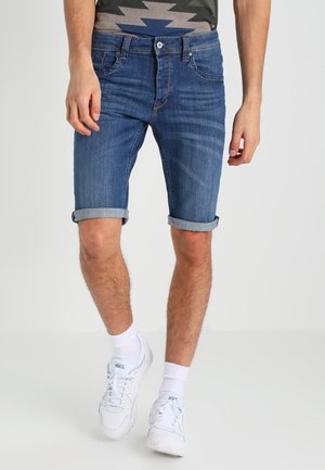 Mand iført op rullede blå denimshorts, hvide ankelstrømper og hvide sneakers, stående mod en ensfarvet lys baggrund.
