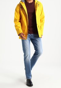 Mann, der eine gelbe Regenjacke, einen kastanienbraunen Pullover, eine hellblaue Jeans und schwarze Stiefel trägt, vor einem weißen Hintergrund läuft.