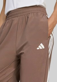 Brown athletic pants with a smocked waistband, side pockets, and white adidas stripes along the sides. Smooth, lightweight fabric texture.