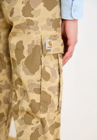 Spodnie cargo Carhartt w kolorze beżowym z kamuflażowym wzorem. Posiadają boczną kieszeń oraz widoczną metkę z logo marki. Tkanina o fakturze z liniami siatki.
