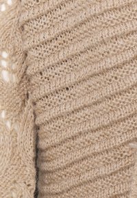 Beige stickad tröja med ett texturerat mönster, som har diagonal ribbning samt mjukt, fluffigt garn och en kabelstickad del.