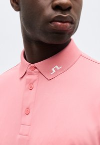 Bărbat purtând un tricou polo roz cu trei nasturi și un mic logo brodat alb pe guler.